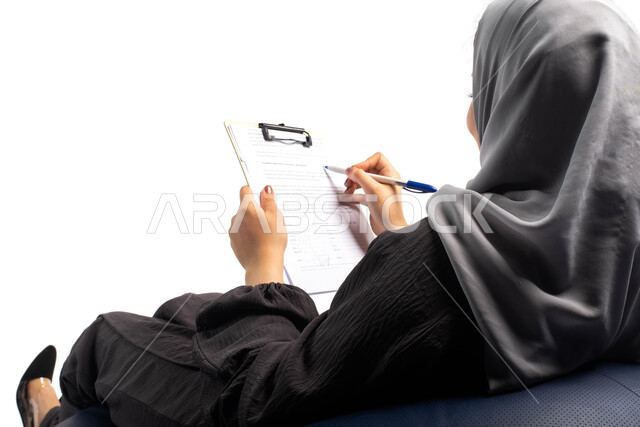 Writing and taking important notes, writing reports and research on paper, a portrait from behind of a young Saudi Arabian Gulf woman sitting on a comfortable chair and holding work papers and files in her hand, white background