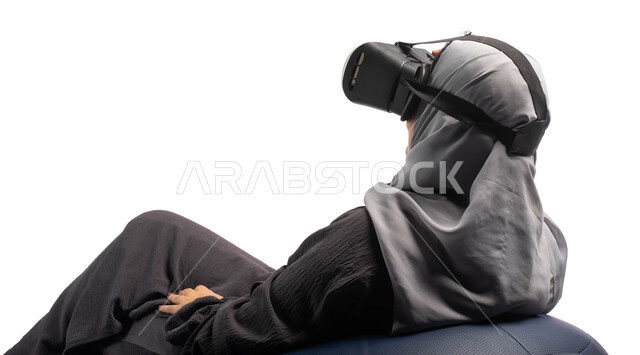 وسائل ترفيهية في وقت الفراغ, الاستمتاع بمشاهدة أفلام الواقع الافتراضي، بورتريه من الخلف لإمرأة عربية خليجية سعودية تجلس على كرسي مريح وترتدي نظارة الواقع الافتراضي ثلاثية الابعاد، قضاء أوقات ممتعة ومسلية بالألعاب الافتراضية، خلفية بيضاء