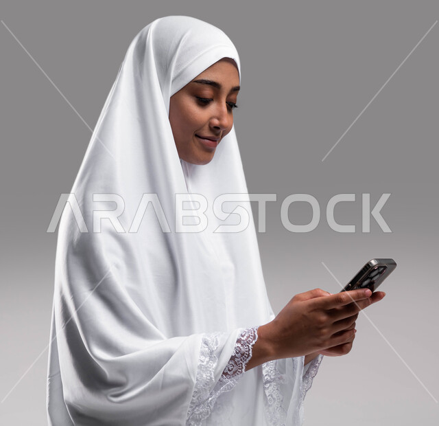 أداء مناسك الحج والعمرة 2023، الركن الخامس من أركان الاسلام, استخدام تطبيقات الهواتف الذكية, بورتريه مقربة لإمراة مسلمة خليجية عربية سعودية ترتدي ملابس الاحرام وتقوم بتصفح الهاتف المحمول، خلفية رمادية