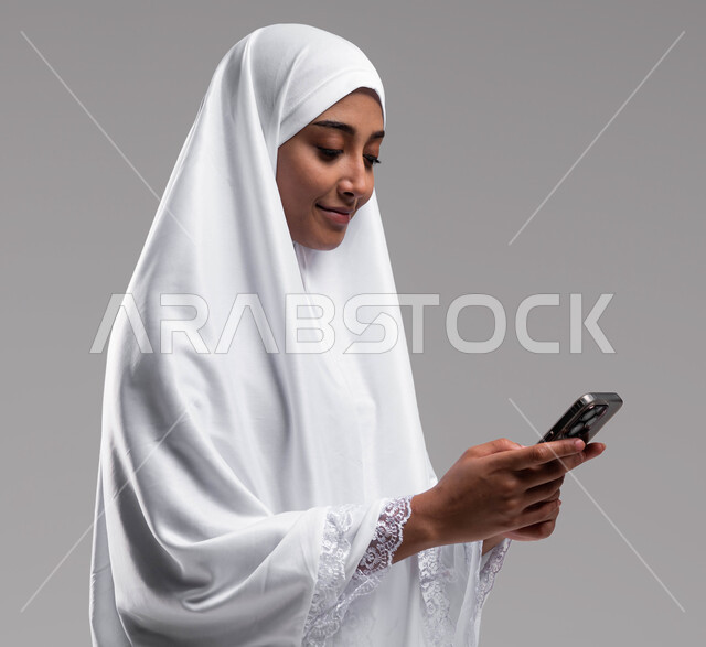 استخدام تطبيقات الهواتف الذكية, بورتريه مقربة لإمراة مسلمة خليجية عربية سعودية ترتدي ملابس الاحرام وتقوم بتصفح الهاتف المحمول، آداء مناسك الحج والعمرة 2023، الركن الخامس من أركان الاسلام, خلفية رمادية