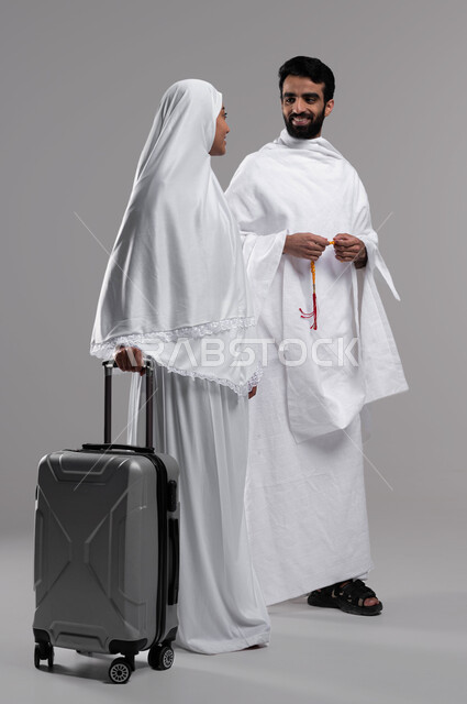 التجهيز والاستعداد للسفر لأداء فريضتي الحج والعمرة 2023 ، بورتريه لزوجان عربيان سعوديان خليجيان مسلمان يرتديان لباس الإحرام، صورة لرجل عربي خليجي سعودي يحمل السبحة الصفراء، امرأة تمسك بيدها حقيبة السفر، خلفية رمادية، إسلاميات وعبادة