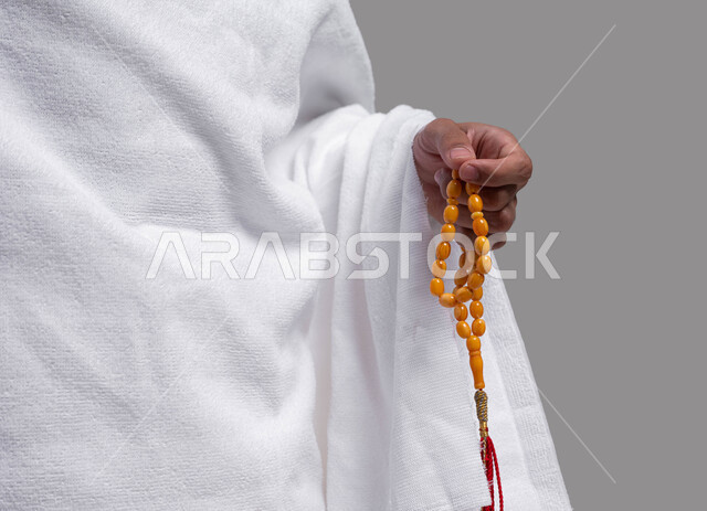 أداء العمرة في مكة المكرمة في شهر رمضان المبارك، بورتريه مقربة لرجل مسلم خليجي عربي سعودي يرتدي ملابس الإحرام ويحمل سبحة صفراء ،  ايماءات وجه تدل على السرور، أداء مناسك الحج والعمرة، إسلاميات وعبادة ، خلفية رمادية.