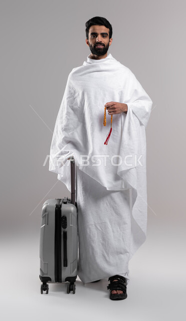 Full body image, wearing ihram clothes, portrait of a Saudi Arabian Gulf Muslim man carrying a rosary and a travel bag, preparing to travel and going to perform Hajj, facial gestures indicating happiness, Hajj season 2023, performing Hajj and Umrah ritual