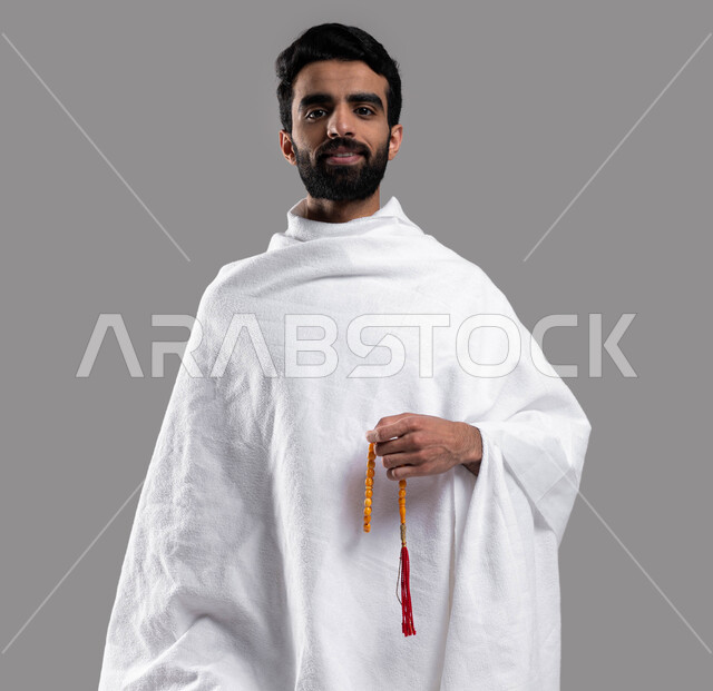 تسبيح وأذكار وأدعية، أداء العمرة في مكة المكرمة في شهر رمضان المبارك، بورتريه مقربة لرجل مسلم خليجي عربي سعودي يرتدي ملابس الإحرام ويحمل سبحة صفراء ،  ايماءات وجه تدل على السرور، أداء مناسك الحج والعمرة، إسلاميات وعبادة ، خلفية رمادية.