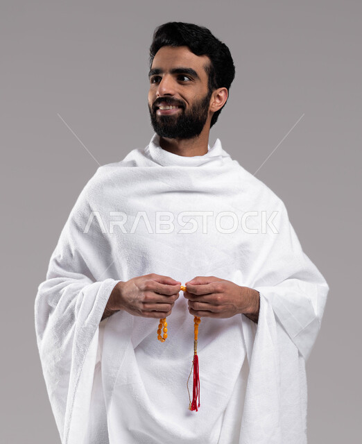 نسبيح وأذكار، أداء العمرة في مكة المكرمة في شهر رمضان المبارك، بورتريه مقربة لرجل مسلم خليجي عربي سعودي يرتدي ملابس الإحرام ويحمل سبحة صفراء ،  ايماءات وجه تدل على السرور، أداء مناسك الحج والعمرة، إسلاميات وعبادة ، خلفية رمادية.