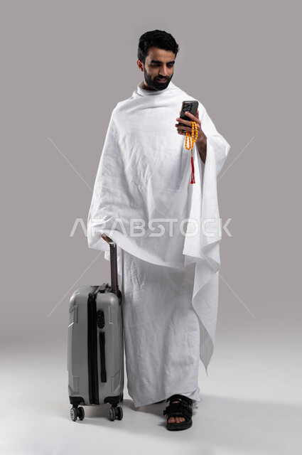 The electronic guide for Hajj and Umrah, preparing to travel and go to perform the Hajj in Makkah Al-Mukarramah, a young Saudi Arabian Gulf Muslim wearing Ihram clothes and holding a travel bag, performing the rituals of Hajj and Umrah, full-length body i