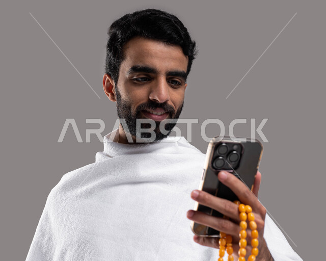 الدعاء عبر الهاتف المحمول ، أداء العمرة في مكة المكرمة، شاب مسلم خليجي عربي سعودي يرتدي ملابس الإحرام ويمسك حقيفة السفر، آداء مناسك الحج والعمرة, صورة الجسم بالطول الكامل،خلفية رمادية.