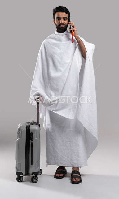 إسلاميات وعبادة، إجراء مكالمة هاتفية عبر الهاتف المحمول، الاستعداد للسفر والذهاب لأداء فريضة الحج في مكة المكرمة، شاب مسلم خليجي عربي سعودي يرتدي ملابس الإحرام ويمسك حقيفة السفر، آداء مناسك الحج والعمرة, صورة الجسم بالطول الكامل،خلفية رمادية.