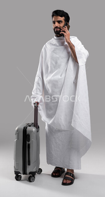  إجراء مكالمة هاتفية عبر الهاتف المحمول، الاستعداد للسفر والذهاب لأداء فريضة الحج في مكة المكرمة، شاب مسلم خليجي عربي سعودي يرتدي ملابس الإحرام ويمسك حقيفة السفر، موسم الحج 2023، آداء مناسك الحج والعمرة, صورة الجسم بالطول الكامل،خلفية رمادية.