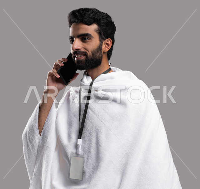 Making a phone call via mobile phone, close-up portrait of a Saudi Arabian Gulf Muslim man wearing ihram clothes and wearing a white card, performing prayers in the mosque, rituals of Hajj and Umrah, the fifth pillar of Islam, Hajj season 2023, gray backg