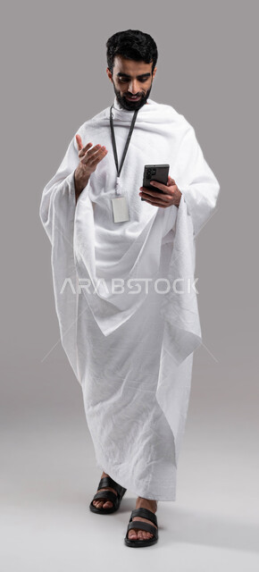 استخدام تطبيق دليل الحاج والمعتمر الالكتروني على الهاتف المحمول, أداء مناسك الحج والعمرة في موسم الحج 2023, بورتريه شاب سعودي خليجي مسلم يرتدي ملابس الإحرام وبطاقة بيضاء تعريفية ويستخدم الجوال ، الركن الخامس من أركان الإسلام ، خلفية رمادية، صورة الجسم بال