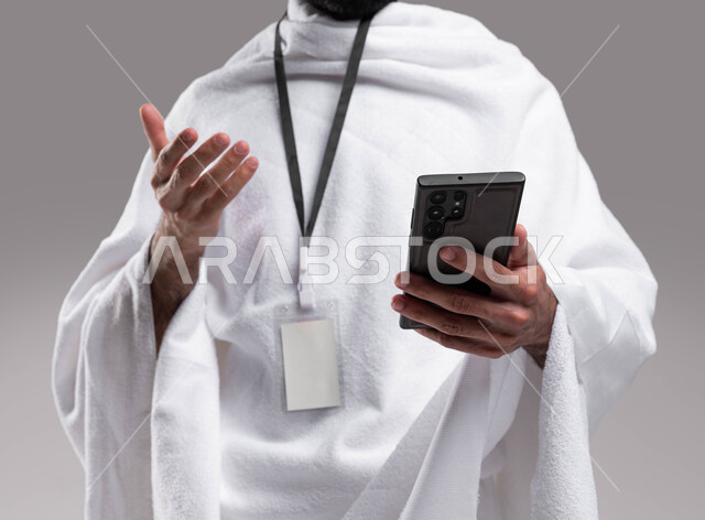 أداء مناسك الحج والعمرة في موسم الحج 2023, استخدام تطبيق دليل الحاج والمعتمر الالكتروني على الهاتف المحمول, بورتريه مقربة لشاب سعودي خليجي مسلم يرتدي ملابس الإحرام وبطاقة بيضاء تعريفية ويستخدم الجوال ، الركن الخامس من أركان الإسلام ، خلفية رمادية، صورة ال