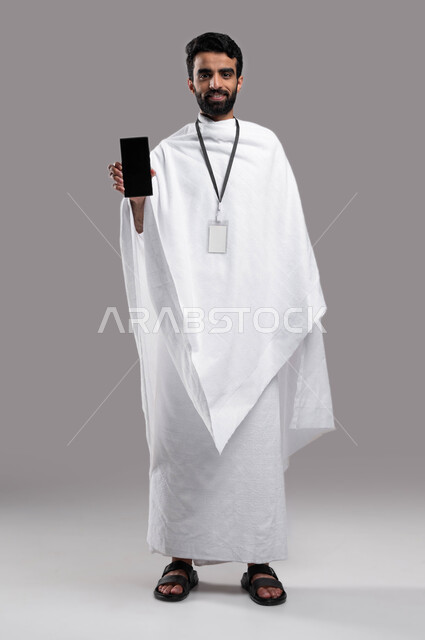Performing the rituals of Hajj and Umrah 2023, facial gestures indicating happiness, drawing closer to God by performing acts of worship, a portrait of a Saudi Arabian Gulf Muslim man wearing ihram clothes and holding a mobile phone with a blank black scr