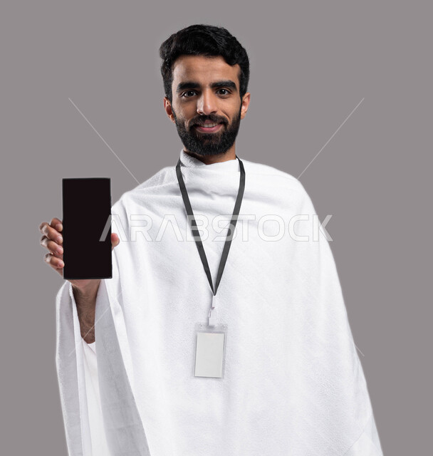 Facial gestures indicating happiness, drawing closer to God by performing acts of worship, a portrait of a Gulf Arab Saudi Muslim man wearing ihram clothes and holding a mobile phone with a blank black screen, Hajj season 2023, performing the rituals of H