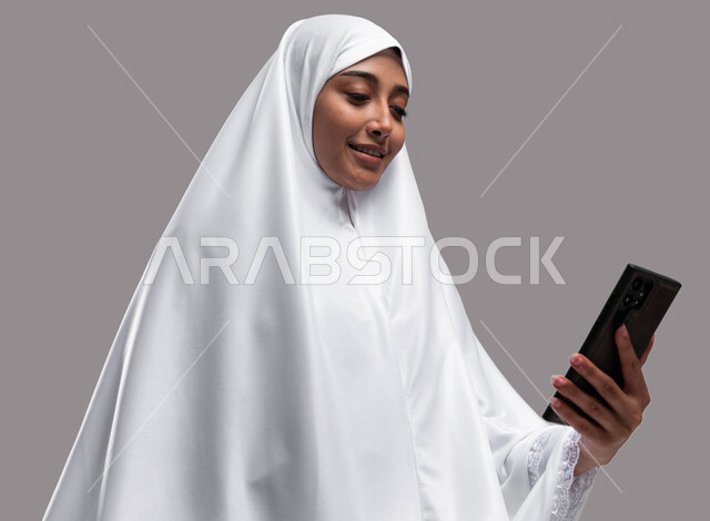 الدعاء عبر الهاتف المحمول, بورتريه لامرأة مسلمة سعودية خليجية عربية ترتدي ملابس الاحرام وتجري مكالمة فيديو, الدليل الالكتروني للحاج و المعتمر, استخدام تطبيقات الجوال للتواصل مع العائلة والأصدقاء, خلفية رمادية
