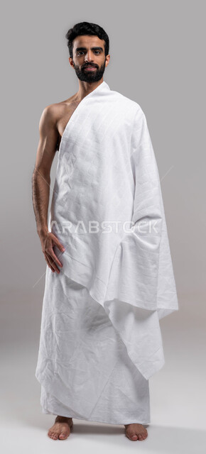 التقرب إلى الله بالأعمال الصالحة، بورتريه لشاب مسلم خليجي عربي سعودي يرتدي ملابس الاحرام، ايماءات تدل على السعادة بآداء مناسك الحج والعمرة، الركن الخامس من أركان الاسلام، خلفية رمادية، صورة الجسم بالطول الكامل. 