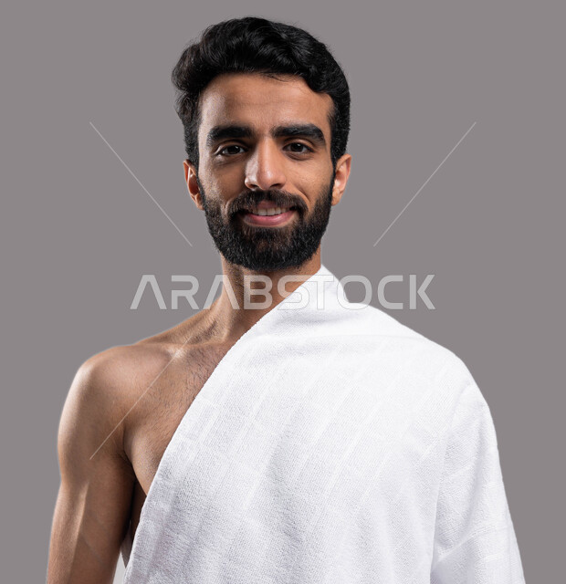 زيارة بيت الله الحرام في مكة المكرمة، التقرب إلى الله بالأعمال الصالحة، بورتريه مقربة لشاب مسلم خليجي عربي سعودي يرتدي ملابس الاحرام، ايماءات تدل على السعادة بآداء مناسك الحج والعمرة، خلفية رمادية. 
