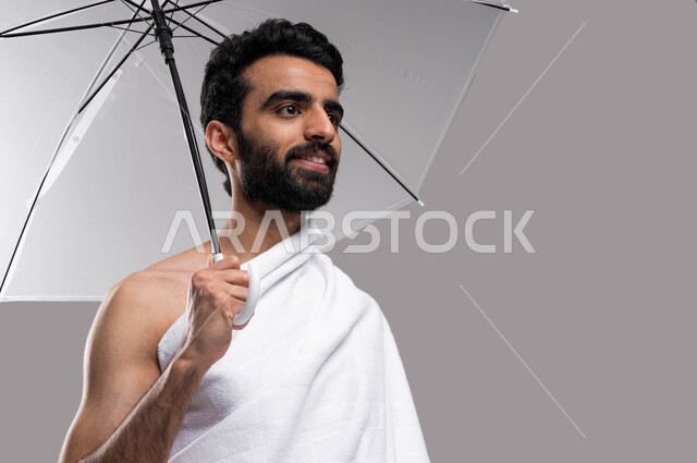 التقرب إلى الله بالأعمال الصالحة، شمسية رمادية صغيرة، بورتريه لشاب مسلم خليجي عربي سعودي يرتدي ملابس الاحرام ويحمل بيده مظلة حامية من المطر، ايماءات تدل على السعادة بآداء مناسك الحج والعمرة، الركن الخامس من أركان الاسلام، خلفية رمادية