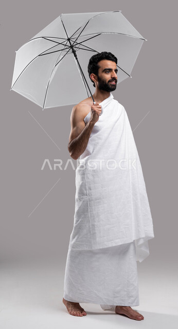  صورة الجسم بالطول الكامل، شمسية رمادية صغيرة، بورتريه لشاب مسلم خليجي عربي سعودي يرتدي ملابس الاحرام ويحمل بيده مظلة حامية من المطر، ايماءات تدل على السعادة بآداء مناسك الحج والعمرة، الركن الخامس من أركان الاسلام، خلفية رمادية