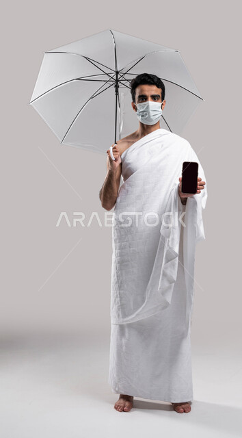 الدعاء عبر الهاتف المحمول، الوقاية من فايروس كورونا، التقرب الى الله بآداء مناسك الحج والعمرة، مظلة حامية من المطر، بورتريه لشاب مسلم خليجي عربي سعودي يرتدي ملابس الاحرام ويحمل بيده شمسية رمادية ويقوم بتصفح الهاتف الجوال، موسم الحج 2023 ، خلفية رمادية