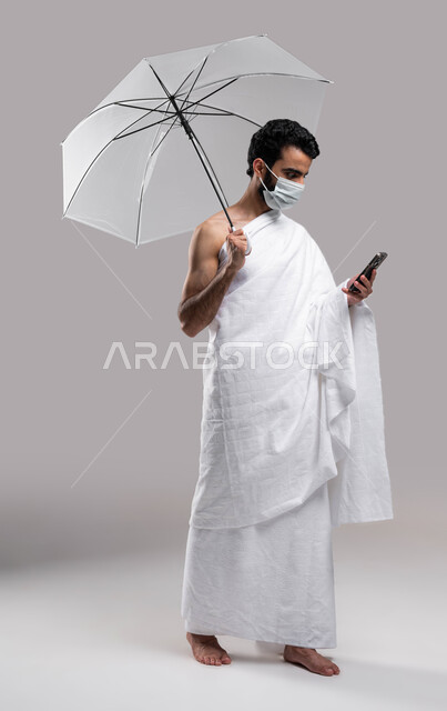 الوقاية من فايروس كورونا، التقرب الى الله بآداء مناسك الحج والعمرة، مظلة حامية من المطر، بورتريه لشاب مسلم خليجي عربي سعودي يرتدي ملابس الاحرام ويحمل بيده شمسية رمادية ويقوم بتصفح الهاتف الجوال، موسم الحج 2023 ، خلفية رمادية