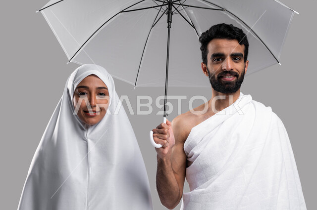 أداء العمرة في شهر رمضان المبارك، إيماءات وجه تدل على السعادة بأداء مناسك الحج والعمرة 2023 ، بورتريه مقربة لزوجين مسلمين خليجيين عربيين سعوديين يرتديان ملابس الاحرام يحملان مظلة حامية من المطر، الركن الخامس من أركان الاسلام، خلفية رمادية. 