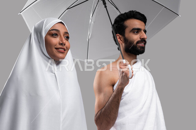 إيماءات وجه تدل على السعادة بأداء مناسك الحج والعمرة 2023 ، بورتريه مقربة لزوجين مسلمين خليجيين عربيين سعوديين يرتديان ملابس الاحرام يحملان مظلة حامية من المطر، الركن الخامس من أركان الاسلام، خلفية رمادية. 