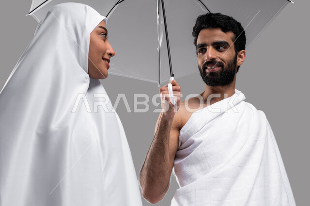 ايماءات تدل على السعادة بآداء مناسك الحج والعمرة، بورتريه مقربة لزوجين مسلمين خليجيين عربيين سعوديين يرتديان ملابس الاحرام ويحملان مظلة حامية من المطر، موسم الحج 2023، الركن الخامس من أركان الاسلام، خلفية رمادية،. 