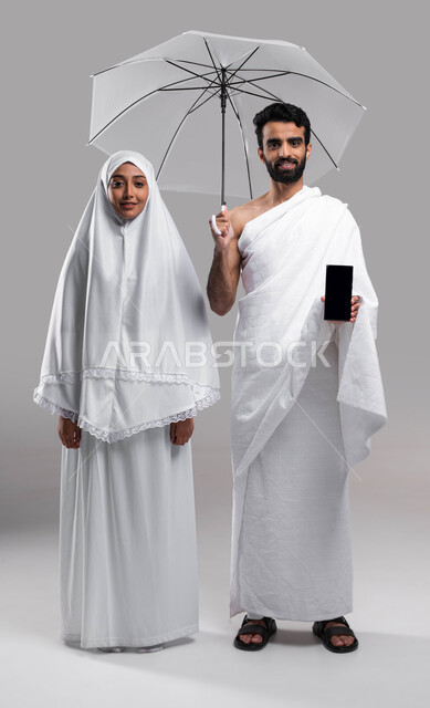 استخدام الهاتف الجوال، بورتريه مقربة لزوجين مسلمين خليجيين عربيين سعوديين يرتديان ملابس الاحرام يحملان مظلة حامية من المطر وهاتف محمول بشاشة سوداء، الركن الخامس من أركان الاسلام، خلفية رمادية، صورة الجسم بالطول الكامل.