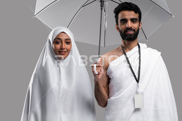 التقرب الى الله و زيارة البيت الحرام، أداء فريضة الحج والعمره في موسم الحج 2023، زوجان عربيان خليحيان سعوديان يرتديان ثياب الاحرام ، العمرة في شهر رمضان المبارك, شاب يحمل بيده مظلة رمادية, خلفية رمادية