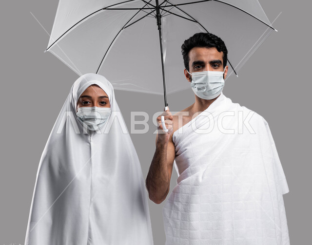 Umrah in the holy month of Ramadan, the concept of charity between spouses, performing the rituals of Hajj and Umrah in the Hajj season 2023, a Saudi Arab Gulf couple wearing Ihram clothes and a protective mask and holding an umbrella in their hands, draw
