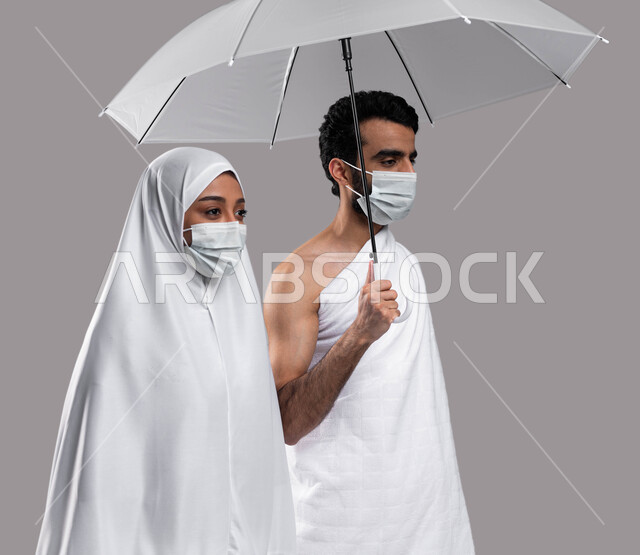 مفهوم الاحسان بين الزوجين، أداء مناسك الحج والعمره في موسم الحج 2023، زوجان عربيان خليحيان سعوديان يرتديان ثياب الاحرام والكمامة الواقية ويحملان بيديهما مظلة، العمرة في شهر رمضان المبارك ، التقرب الى الله بالأعمال الصالحة, خلفية رمادية