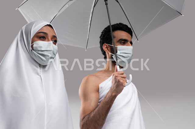 مفهوم الاحسان بين الزوجين، أداء مناسك الحج والعمره في موسم الحج 2023، زوجان عربيان خليحيان سعوديان يرتديان ثياب الاحرام والكمامة الواقية ويحملان بيديهما مظلة، العمرة في شهر رمضان المبارك ، التقرب الى الله بالأعمال الصالحة, خلفية رمادية