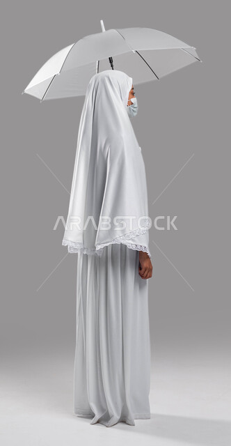 الالتزام بالإجراءات الوقائية لكورونا في موسم الحج 2023، زيارة مكة المكرمة لأداء مناسك الحج والعمرة, اسلاميات وعبادة, بورتريه من الجانب شابة مسلمة خليجية عربية سعودية ترتدي ملابس الاحرام وتحمل بيدها مظلة حامية من المطر، صورة الجسم بالطول الكامل, خلفية رماد