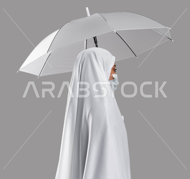 زيارة مكة المكرمة لأداء مناسك الحج والعمرة, اسلاميات وعبادة, بورتريه من الجانب شابة مسلمة خليجية عربية سعودية ترتدي ملابس الاحرام وتحمل بيدها مظلة حامية من المطر، الالتزام بالإجراءات الوقائية لكورونا في موسم الحج 2023، خلفية رمادية
