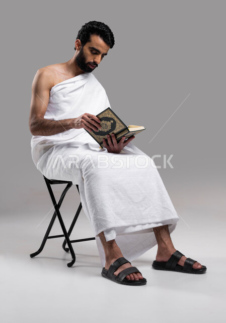 اسلاميات وعبادة, الاجتهاد في الطاعات في الأشهر المباركة, التقرب الى الله بقراءة القرآن, بورتريه لشاب عربي سعودي خليجي يرتدي لباس الاحرام يجلس على كرسي و يحمل بيده كتاب القرآن الكريم ، اداء مناسك الحج والعمرة 2023، خلفية رمادية