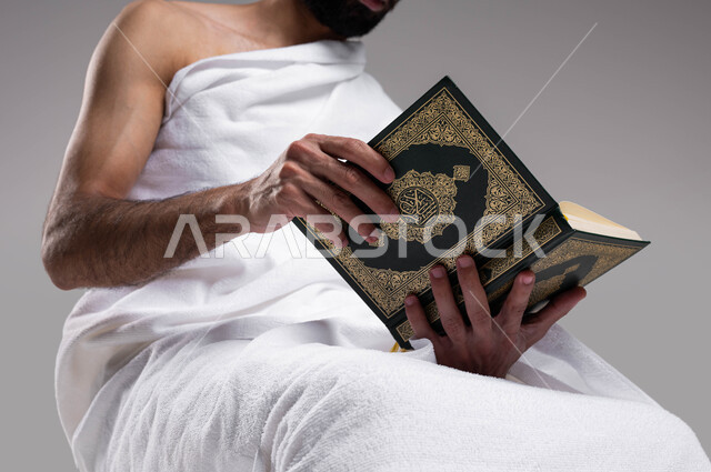 اسلاميات وعبادة, الاجتهاد في الطاعات في الأشهر المباركة, التقرب الى الله بقراءة القرآن, بورتريه لشاب عربي سعودي خليجي يرتدي لباس الاحرام و يحمل بيده كتاب القرآن الكريم ، اداء مناسك الحج والعمرة 2023، خلفية رمادية