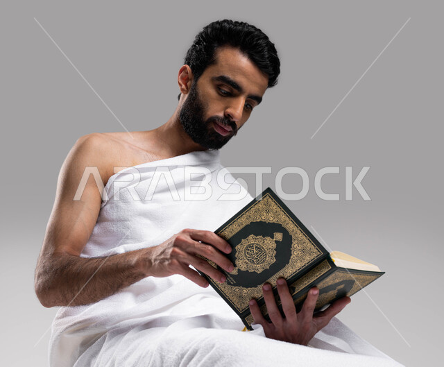 Diligence in obedience in the blessed months, Islam and worship, getting closer to God by reading the Qur’an, a portrait of a young Saudi Arab Gulf man wearing the Ihram garment and holding the Holy Qur’an book in his hand, performing the rituals of Hajj 