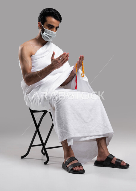 ارتداء الكمامة الواقية، أدعية وأذكار اسلامية , التقرب الى الله بالصلاة والدعاء، آداء مناسك الحج والعمرة في موسم 2023، بورتريه لشاب مسلم عربي سعودي خليجي يجلس على كرسي و يرتدي ملابس الاحرام ويرفع يديه للدعاء ويحمل بيده سبحة، خلفية رمادية