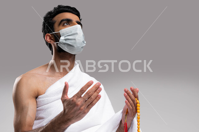 أدعية وأذكار اسلامية , التقرب الى الله بالصلاة والدعاء والعبادة، آداء مناسك الحج والعمرة في موسم 2023، بورتريه لشاب مسلم عربي سعودي خليجي يرتدي ملابس الاحرام ويرفع يديه للدعاء ويحمل بيده سبحة، ارتداء الكمامة الواقية، خلفية رمادية