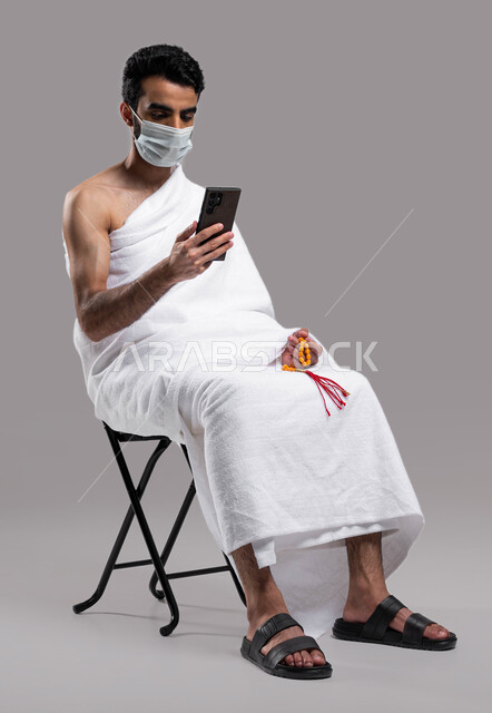 استخدام تطبيقات الجوال للتواصل مع العائلة والأصدقاء, الالتزام بالإجراءات الاحترازية لكورونا, الدعاء عبر الهاتف المحمول, بورتريه لرجل مسلم سعودي خليجي عربي يرتدي ملابس الاحرام والكمامة ويجري مكالمة فيديو, الدليل الالكتروني للحاج و المعتمر, خلفية رمادية