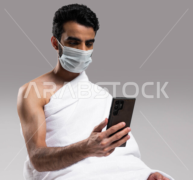 استخدام تطبيقات الجوال للتواصل مع العائلة والأصدقاء, الالتزام بالإجراءات الاحترازية لكورونا, الدعاء عبر الهاتف المحمول, بورتريه لرجل مسلم سعودي خليجي عربي يرتدي ملابس الاحرام والكمامة ويجري مكالمة فيديو, الدليل الالكتروني للحاج و المعتمر, خلفية رمادية