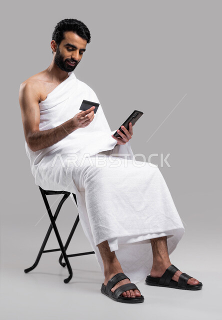 الحجز الالكتروني في حملات الحج والعمرة, الشراء والدفع عن طريق بطاقة الصراف الإئتمانية, بورتريه لشاب مسلم عربي سعودي خليجي يرتدي زي الاحرام ويستخدم الهاتف الجوال وكرت البنك المصرفي, حجوزات الاونلاين السريعة, خلفية رمادية