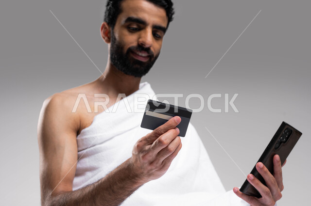 الحجز الالكتروني في حملات الحج والعمرة, الشراء والدفع عن طريق بطاقة الصراف الإئتمانية, بورتريه لشاب مسلم عربي سعودي خليجي يرتدي زي الاحرام ويستخدم الهاتف الجوال وكرت البنك المصرفي, حجوزات الاونلاين السريعة, خلفية رمادية