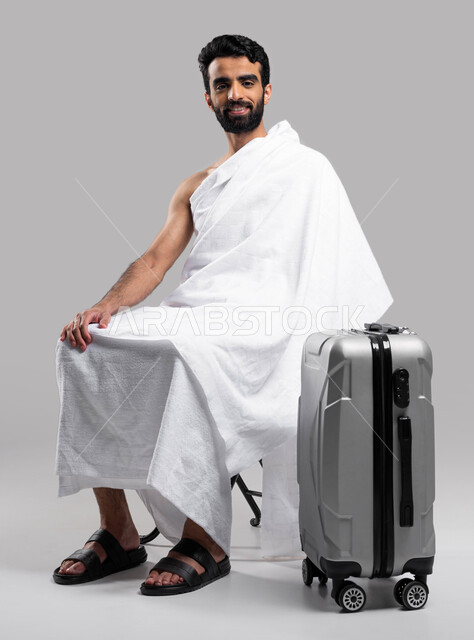 Islamism and worship, preparing and preparing to travel to perform the rituals of Hajj and Umrah 2023, drawing closer to God by performing acts of worship, a portrait of a young Saudi Arabian Gulf Muslim wearing Ihram clothes sitting on a chair with a tra