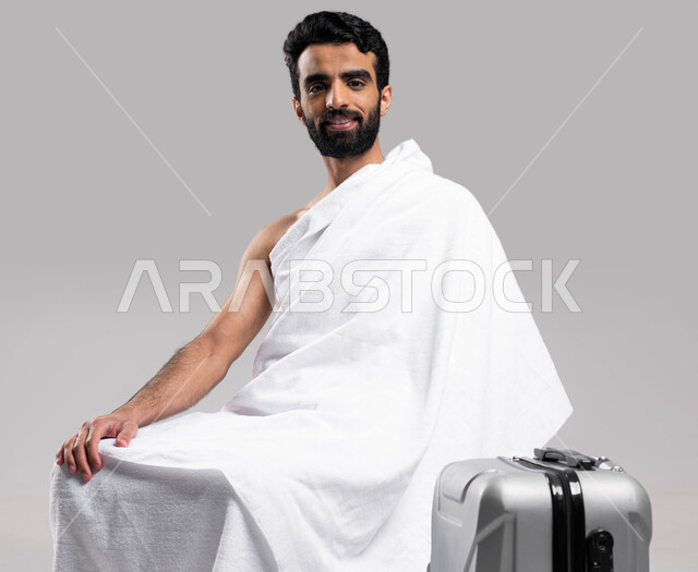 Islamism and worship, preparing and preparing to travel to perform the rituals of Hajj and Umrah 2023, drawing closer to God by performing acts of worship, a portrait of a young Saudi Arabian Gulf Muslim wearing Ihram clothes sitting on a chair with a tra