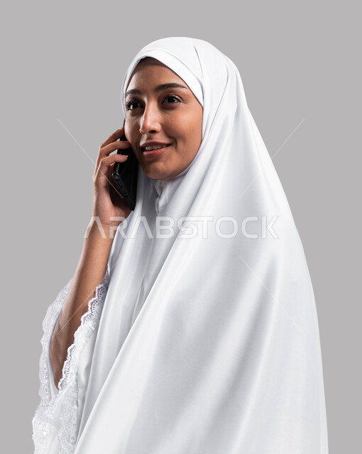 إيماءات وتعابير وجه تدل على السعادة بالتواصل الاجتماعي والعلاقات الاجتماعية، آداء مناسك الحج والعمرة 2023، استخدام الهاتف المحمول، بورتريه صورة جانبية لإمراة مسلمة خليجية عربية سعودية ترتدي ملابس الاحرام و تجري مكالمة هاتفية، خلفية رمادية