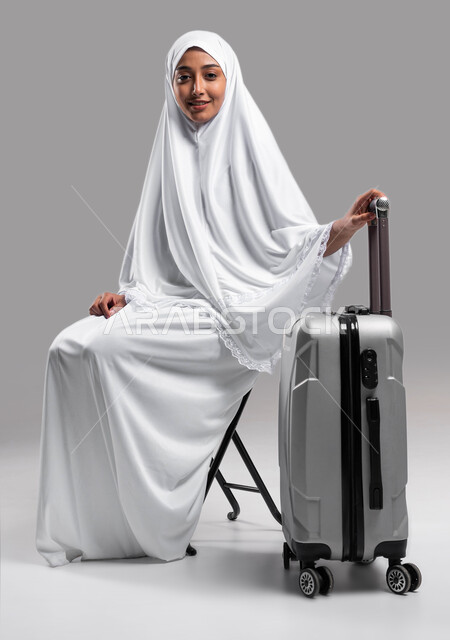 التأهب والاستعداد للسفر لأداء مناسك الحج والعمرة 2023, زيارة بيت الله الحرام في مكة المكرمة, التقرب الى الله بأداء الطاعات, بورتريه لإمراة مسلمة خليجية عربية سعودية ترتدي ملابس الاحرام تجلس على الكرسي وتمسك بيدها حقيبة السفر ، خلفية رمادية
