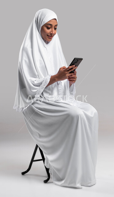 Gestures and facial expressions indicating happiness by making a video call, using mobile phone applications, a portrait of a Saudi Arabian Gulf Muslim woman wearing ihram clothes sitting on a chair and holding a mobile phone in her hand, the electronic g