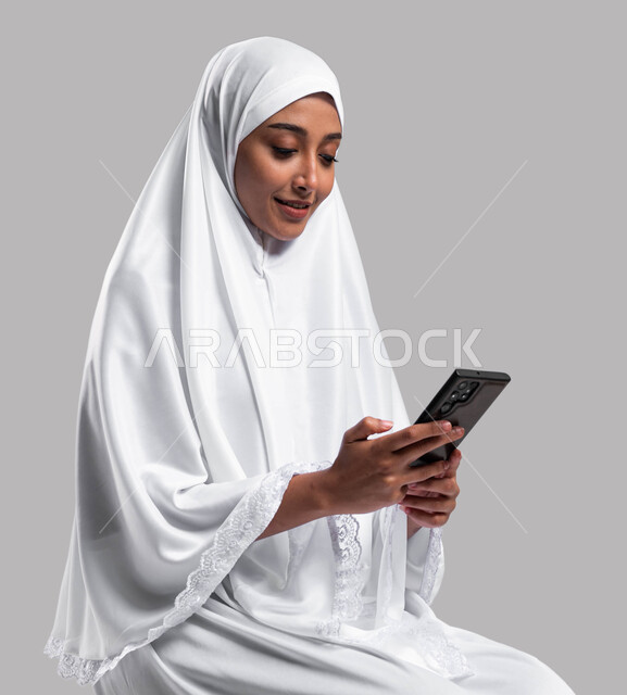 الدليل الالكتروني للحاج والمعتمر, استخدام تطبيقات الهاتف المحمول, بورتريه لإمرأة مسلمة عربية سعودية خليجية ترتدي ملابس الاحرام وتحمل بيدها الهاتف المحمول, إيماءات وتعابير وجه تدل على السعادة بإجراء مكالمة فيديو, موسم الحج2023, خلفية رمادية